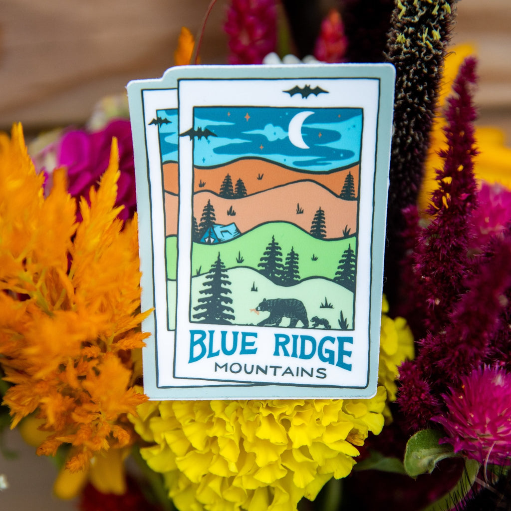 A vinyl sticker with a Blue Ridge Mountains design, featuring a moonlit landscape with trees and a cabin under the moon and stars.