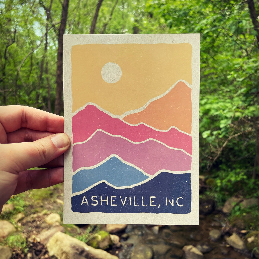 A postcard depicting a stylized mountain range in colors from pink to blue, with the text 'Asheville, NC' at the bottom, held in a hand with a blurred natural background.