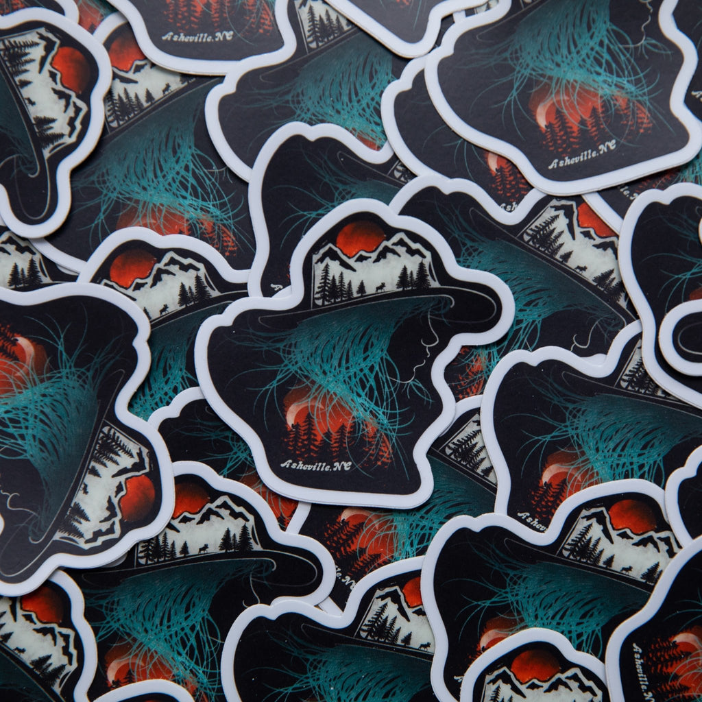 Vinyl sticker shaped like a hat with a mountain landscape and aurora-like teal glow, over a pile of identical stickers, with "Asheville NC" printed in white.

