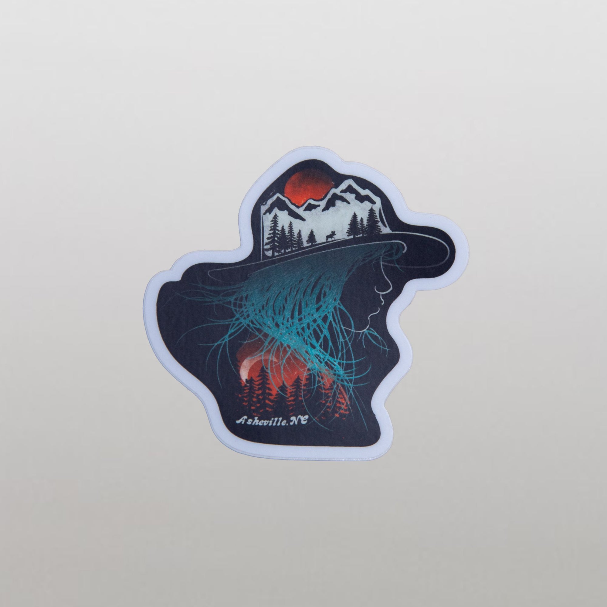 Vinyl sticker shaped like a hat with a mountain landscape and aurora-like teal glow, over a pile of identical stickers, with "Asheville NC" printed in white.

