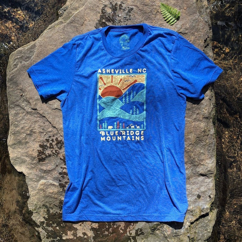 A blue colored t-shirt with a graphic print of mountains and the text 'Asheville, NC' placed on a rock.