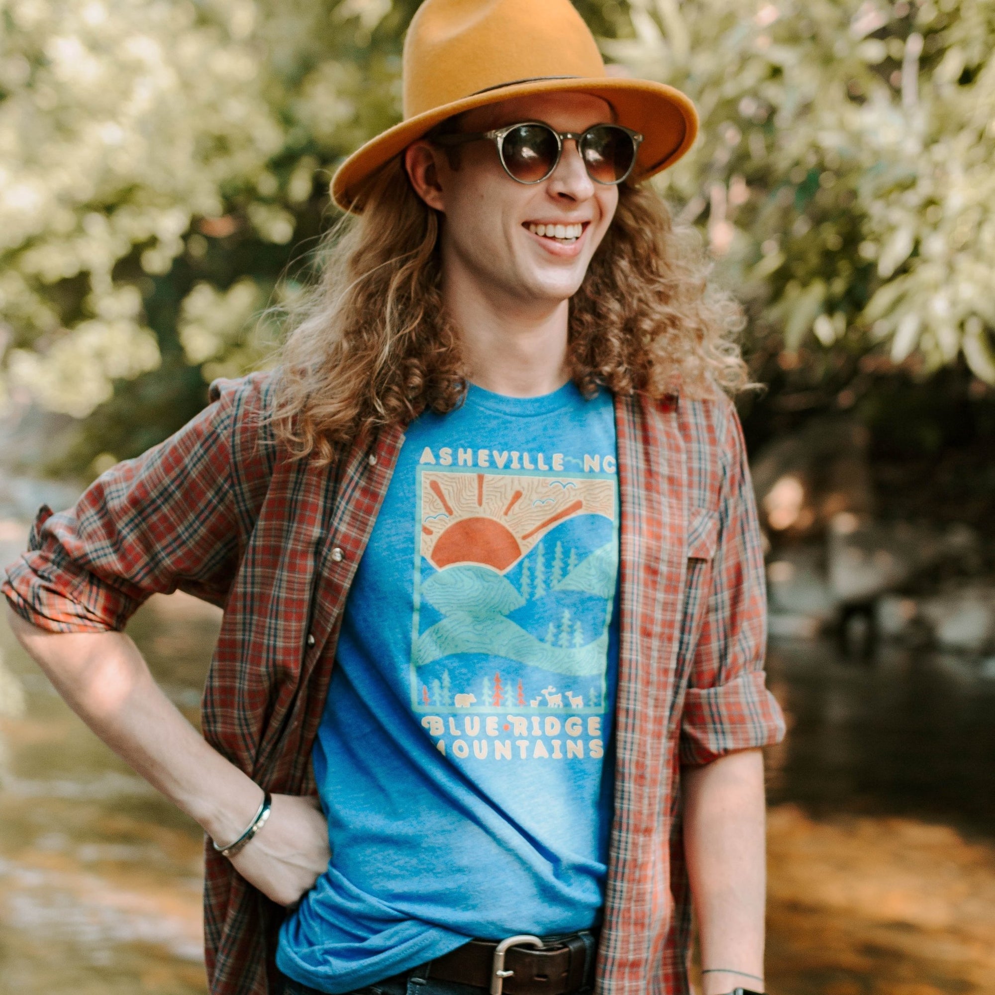 Model wearing the shirt with sunglasses and a hat, standing in the woods with sunlight behind.

