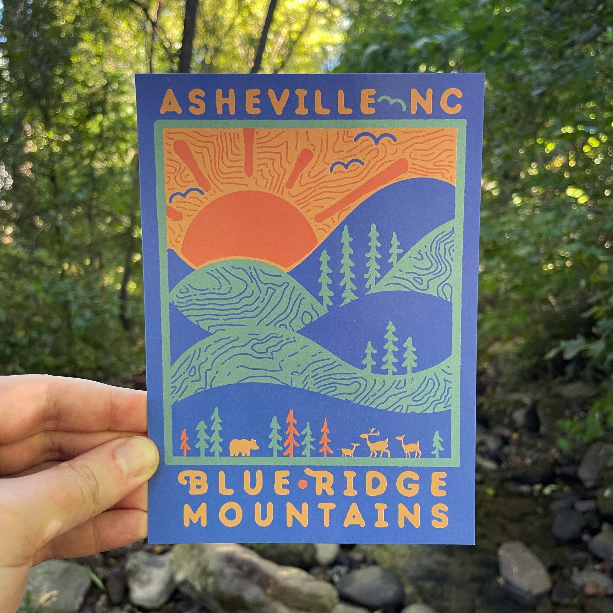 A postcard depicting the Blue Ridge Mountains with a sunburst and trees, held in a hand with a forest background.