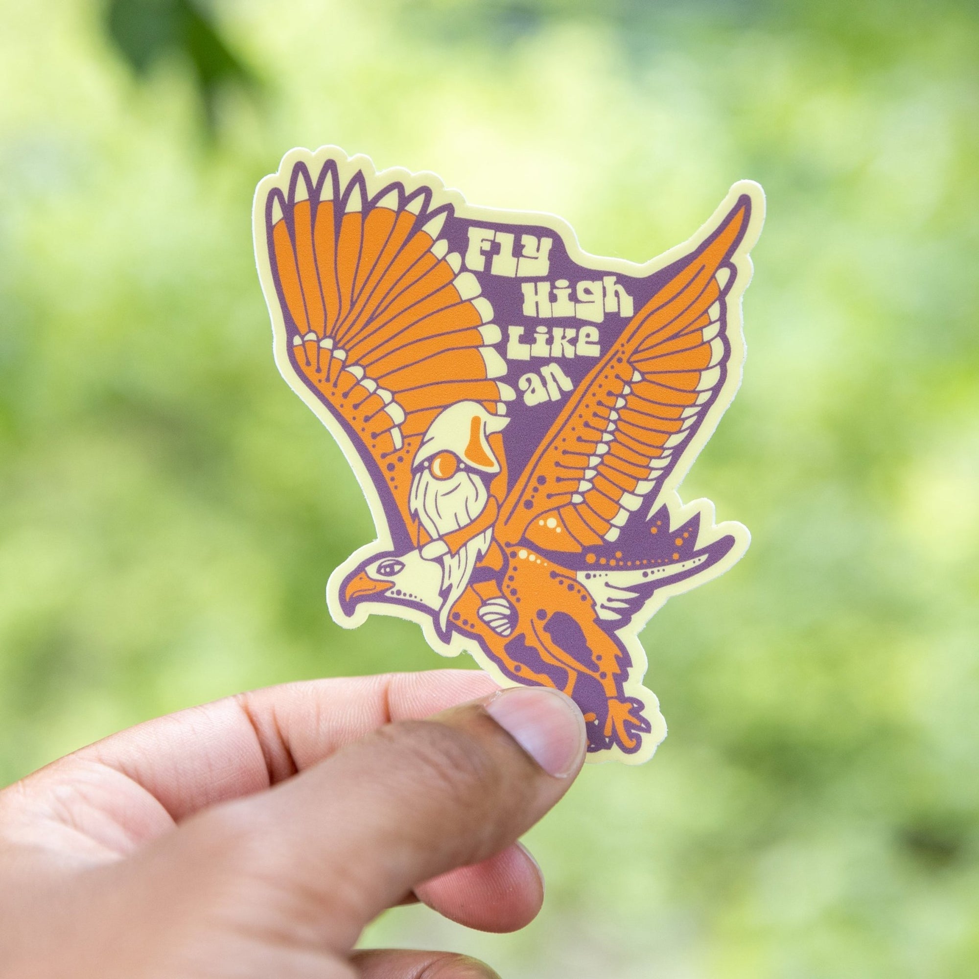 Hand holding vinyl sticker of gnome flying on eagle with “Fly High Like An” text

