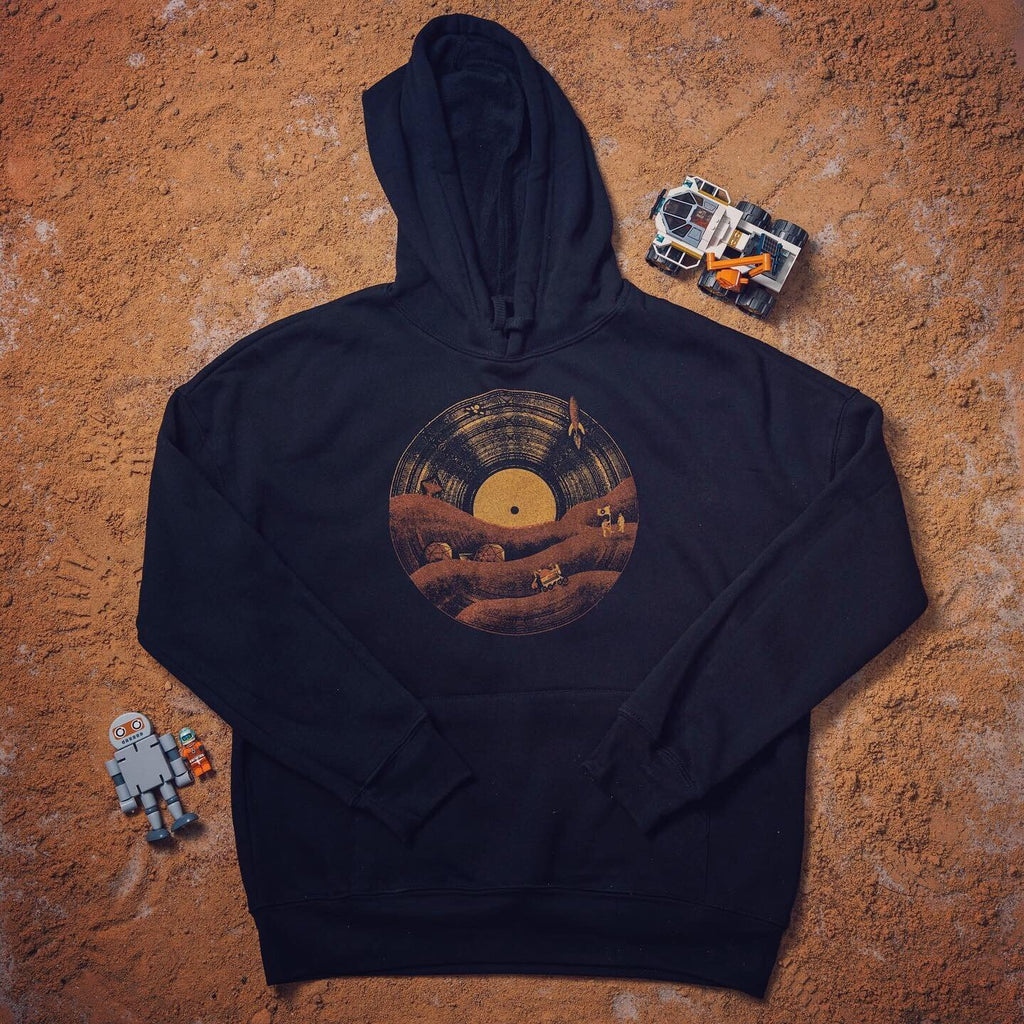 Black hoodie with a vinyl record and Mars rover illustration, laid flat on a red Martian-style surface with a robot and toy rover beside it.

