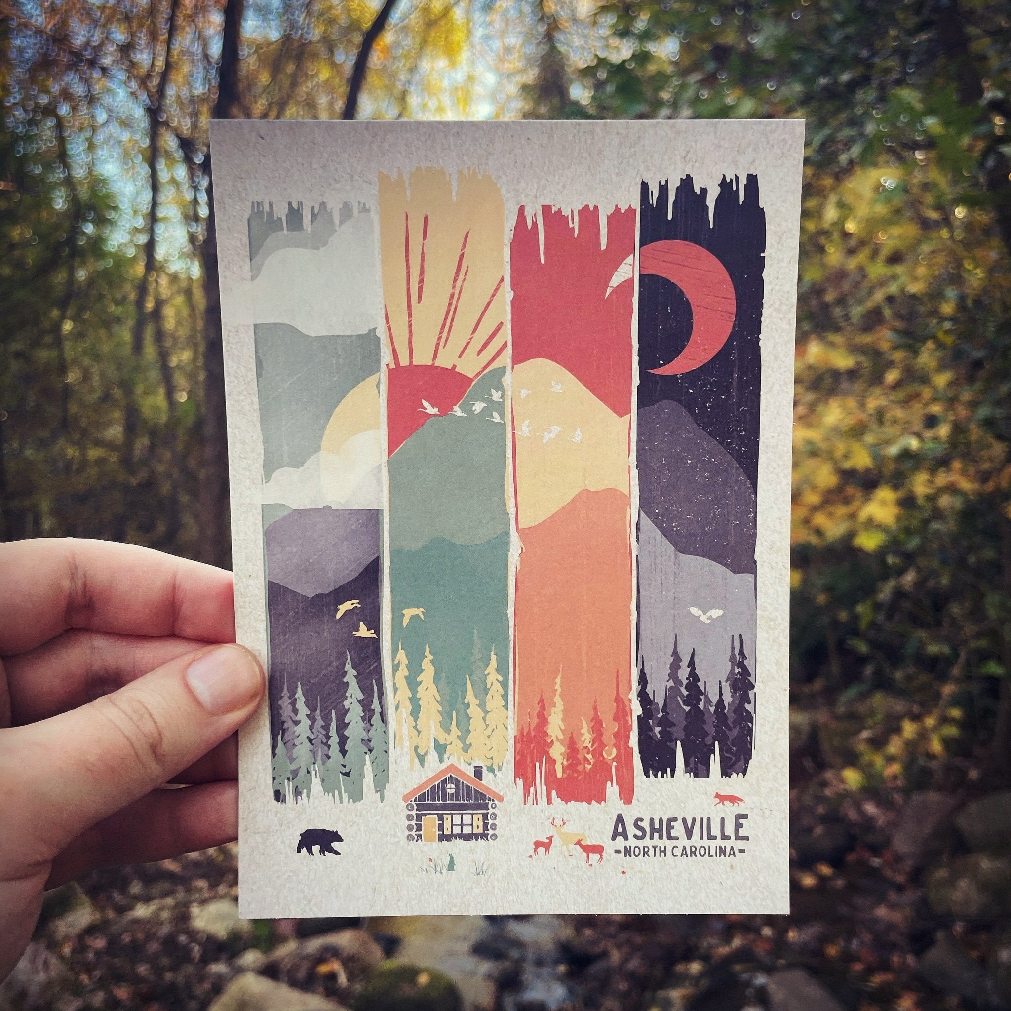 A postcard depicting a stylized mountain scene with a sun and moon, held in a hand, with 'Asheville, North Carolina' printed at the bottom.