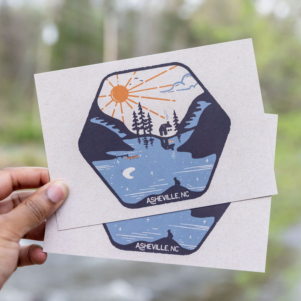 A postcard with a graphic design that includes a stylized sun, trees, and a cabin under the moonlight, with 'Ashville, NC' printed at the bottom. The postcard has a glossy finish.