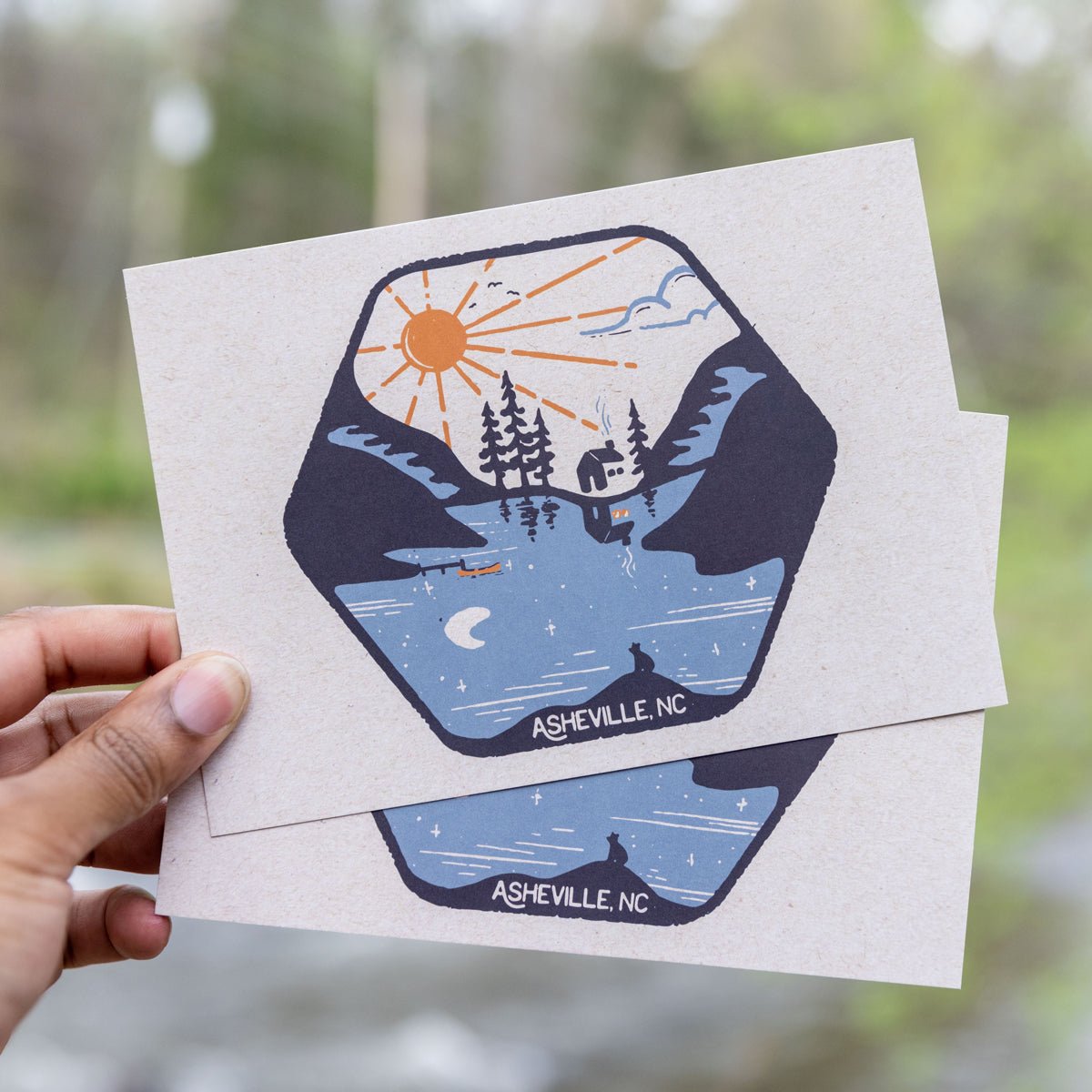 A postcard with a graphic design that includes a stylized sun, trees, and a cabin under the moonlight, with 'Ashville, NC' printed at the bottom. The postcard has a glossy finish.