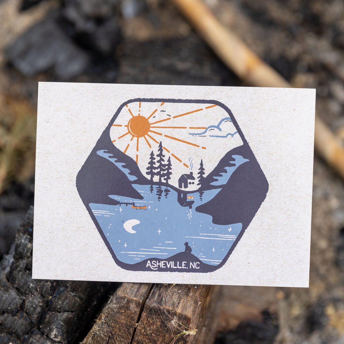 Postcard featuring a mirrored mountain and lake scene with sun and moon, propped upright on a rock with a blurred natural background.