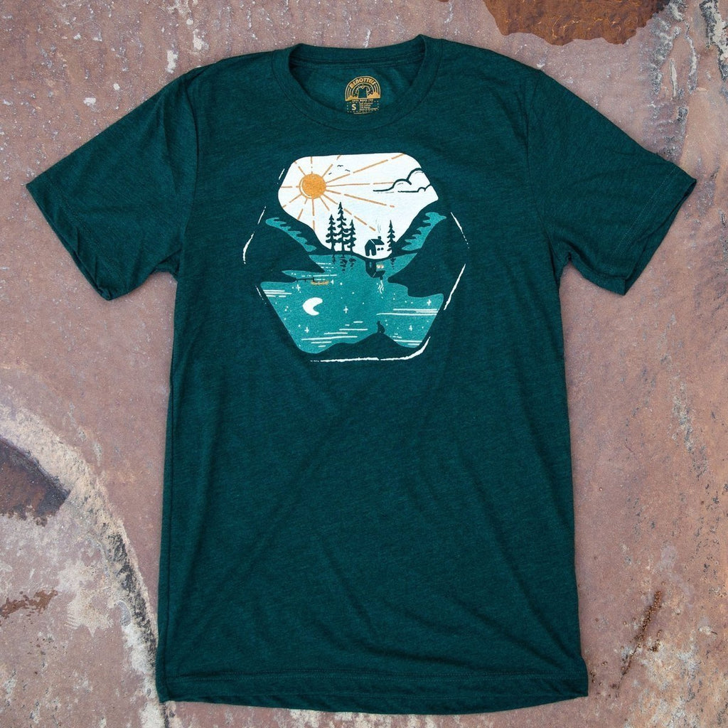 A green crew neck t-shirt with a graphic print of a nature scene including trees and a sun, styled on a rustic background.