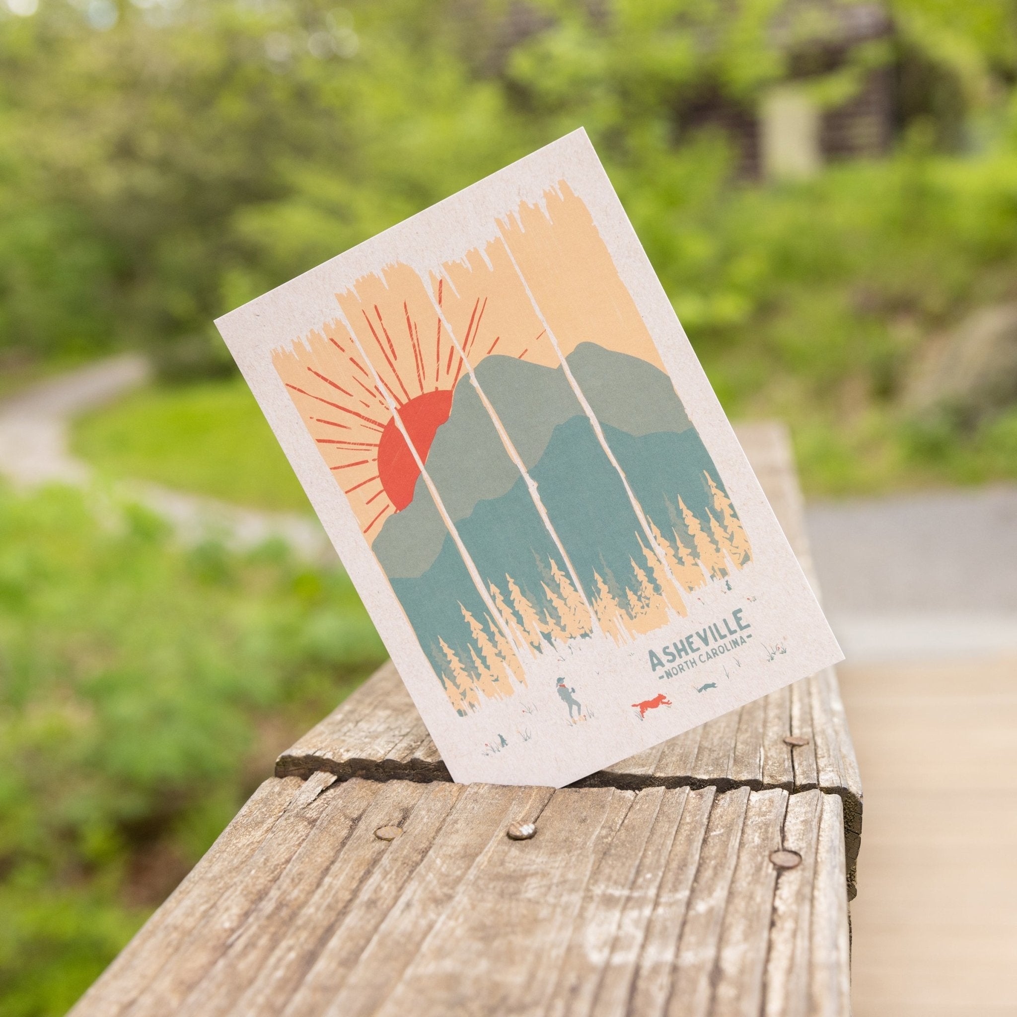Postcard leaning against a wooden railing, featuring a stylized sun setting behind mountain peaks and pine trees, with Asheville, NC printed at the bottom.


