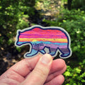 Hand holding a vibrant bear-shaped vinyl sticker with a painted sunset inside the silhouette and "Asheville NC" on the bear’s side, with a forest stream in the background.

