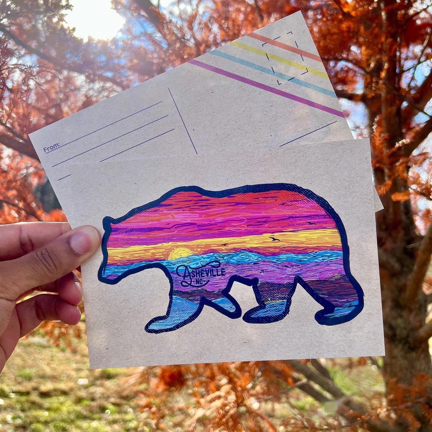 Hand holding postcard with a bear silhouette filled with vibrant sunset colors and Asheville, NC text

