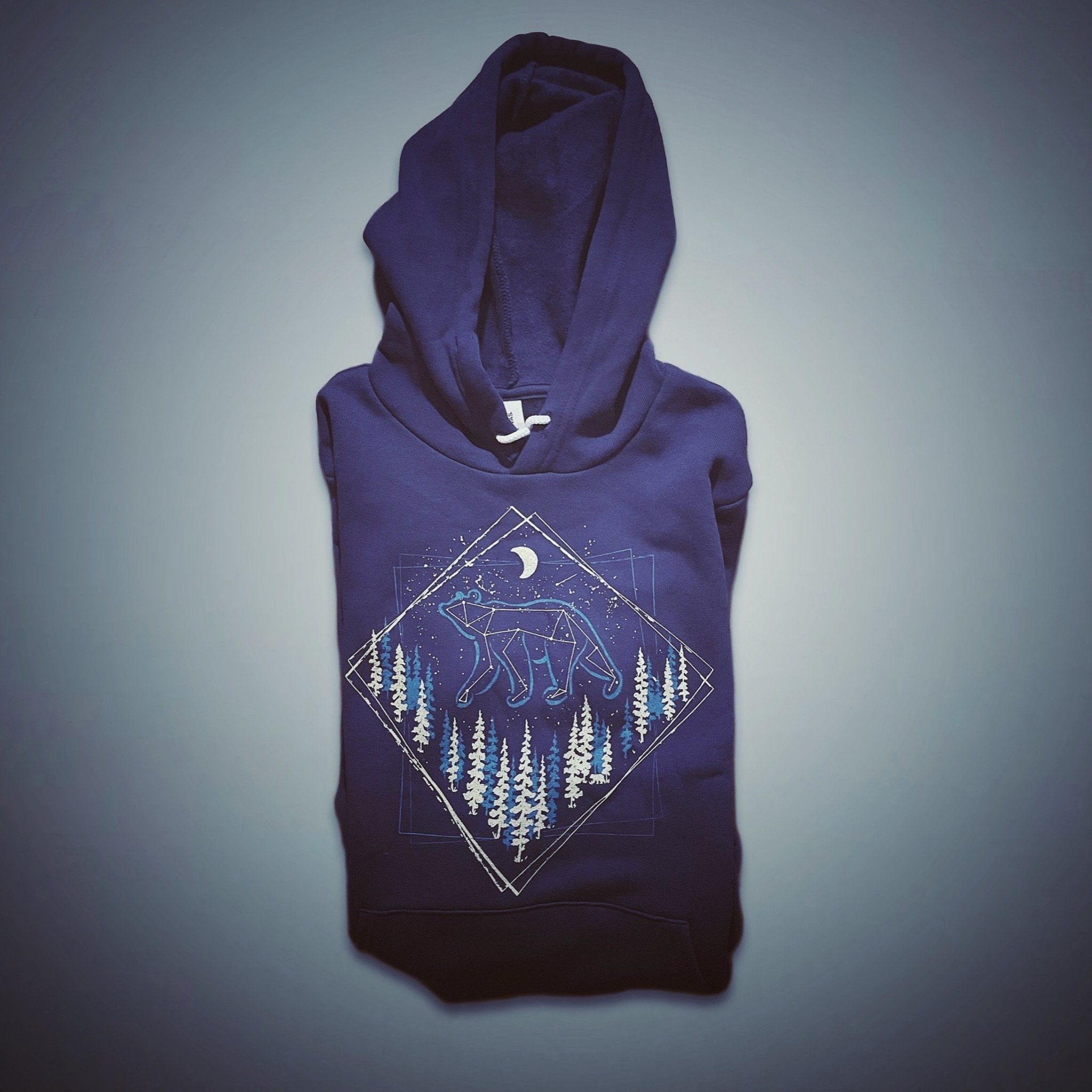 Folded navy hoodie featuring a hand-printed design of a celestial bear walking through pine trees under a moonlit sky, laid out on a soft gradient background.

