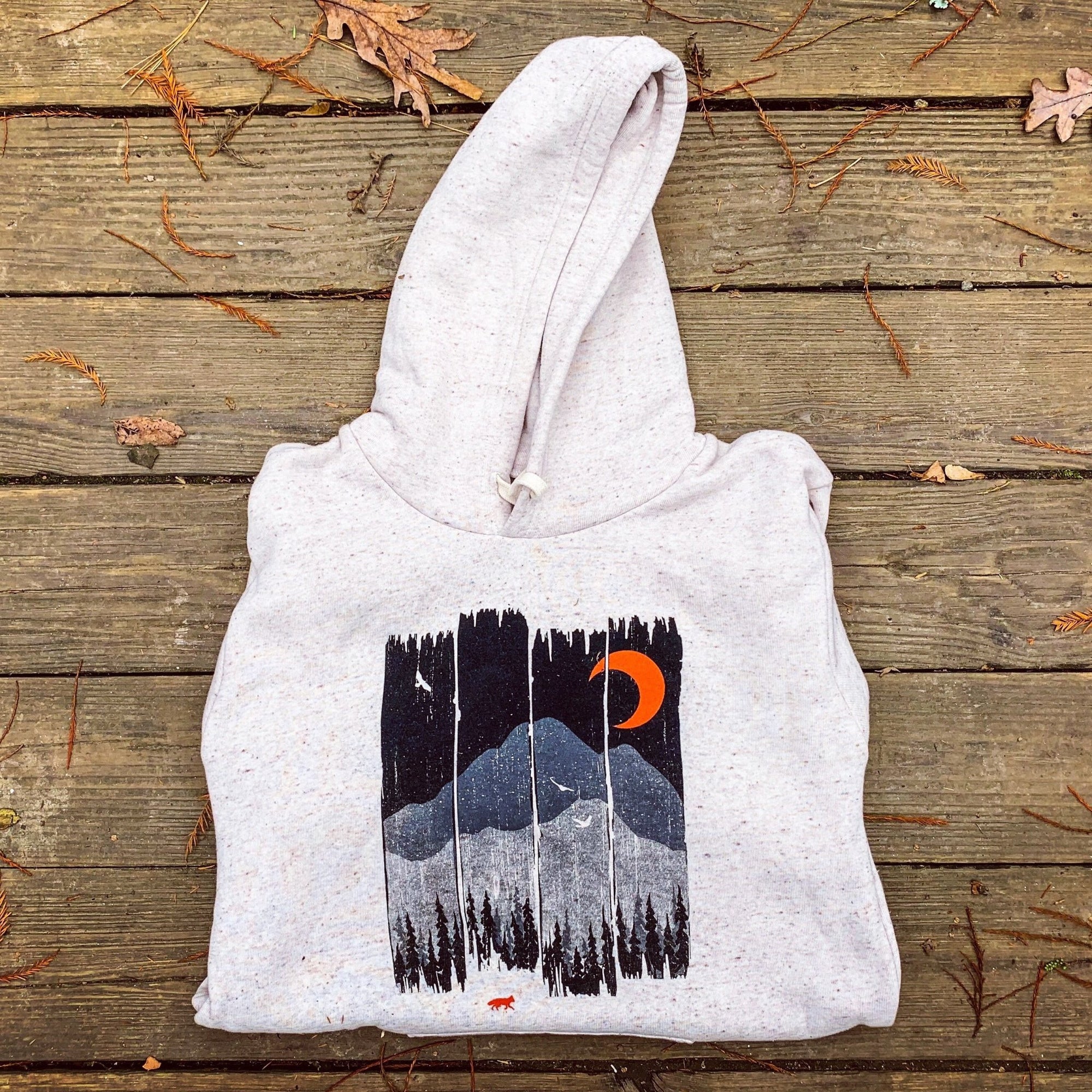 Folded hoodie featuring a hand-screened design of a mountain range at night with a burnt orange moon, displayed on a rustic wooden background.


