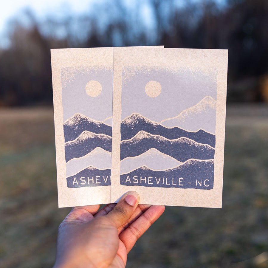 Hand holding two identical Winter Range postcards with snowy mountain design and Asheville, NC text

